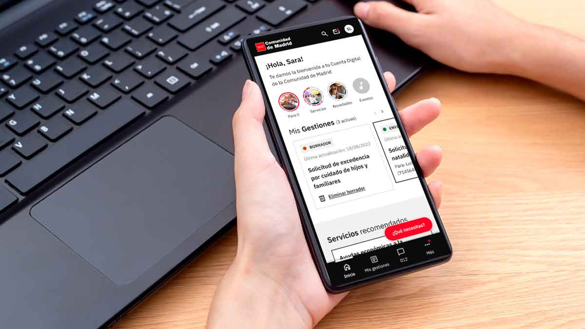 Persona sosteniendo un smartphone con la app Cuenta Digital de la Comunidad de Madrid abierta, gestionando trámites públicos junto a un ordenador portátil
