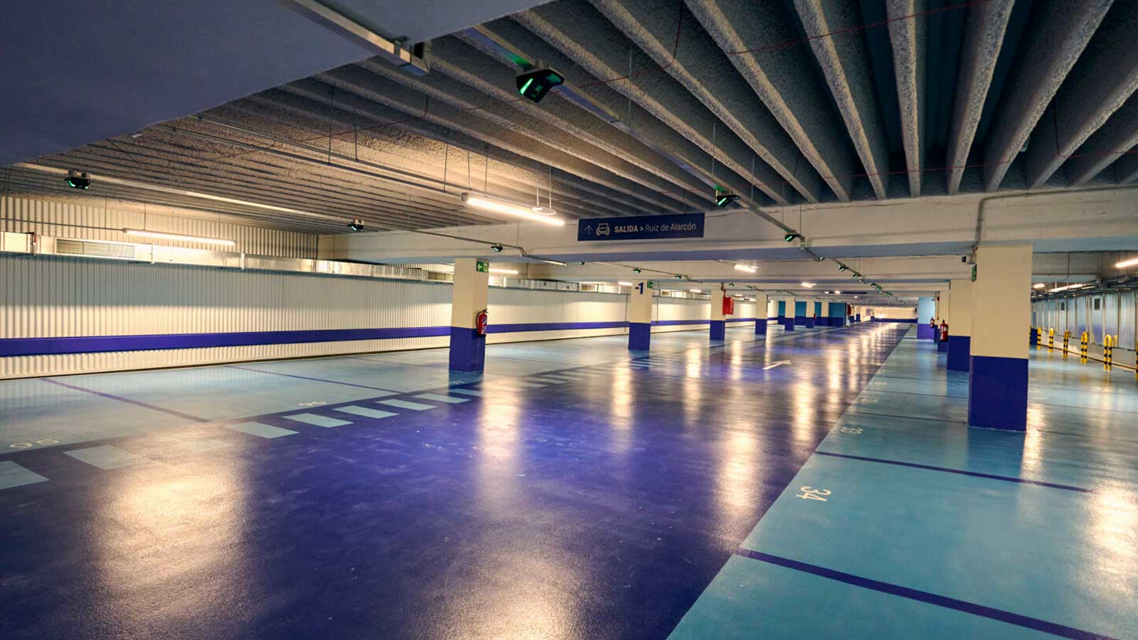 Interior de un aparcamiento subterráneo reformado en Madrid, con plazas de estacionamiento, suelo azul y nueva iluminación.