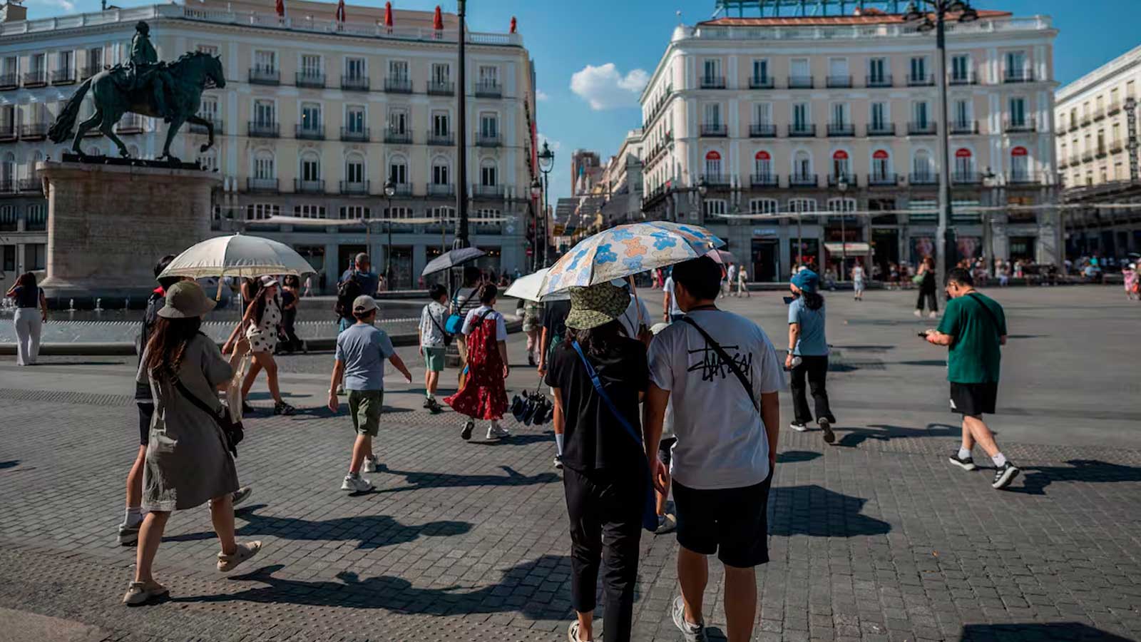 Personas paseando por la Puerta del Sol de Madrid bajo un cielo despejado y protegidas del sol con sombrillas.