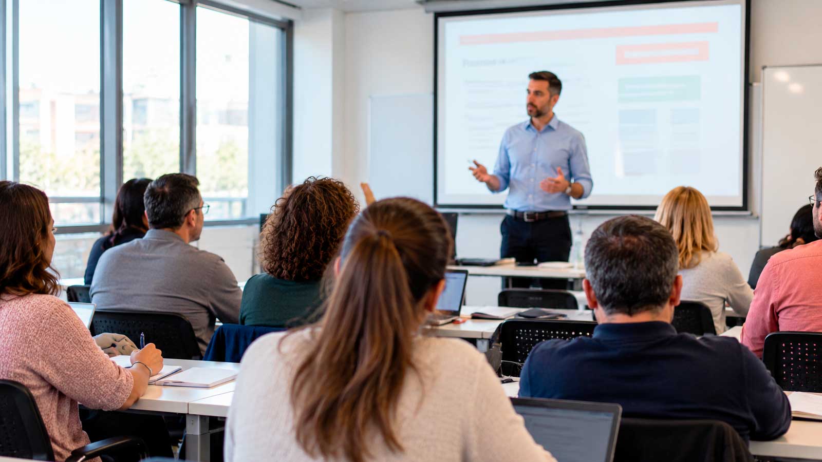 Alumnos adultos asisten a un curso de formación en un aula con un profesor impartiendo clase.