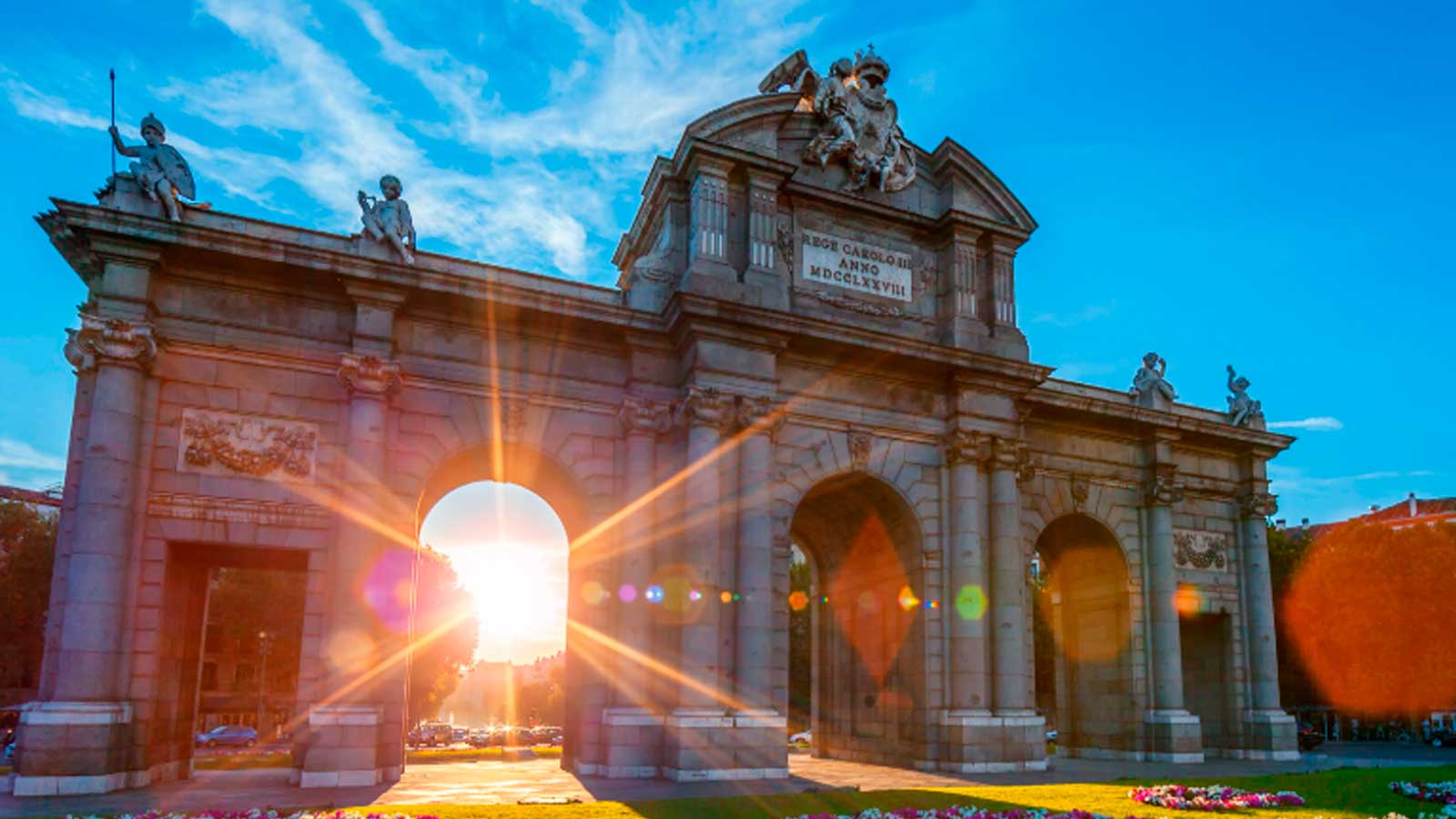 Puerta de Alcalá de Madrid iluminada por el sol bajo un cielo azul con algunas nubes.