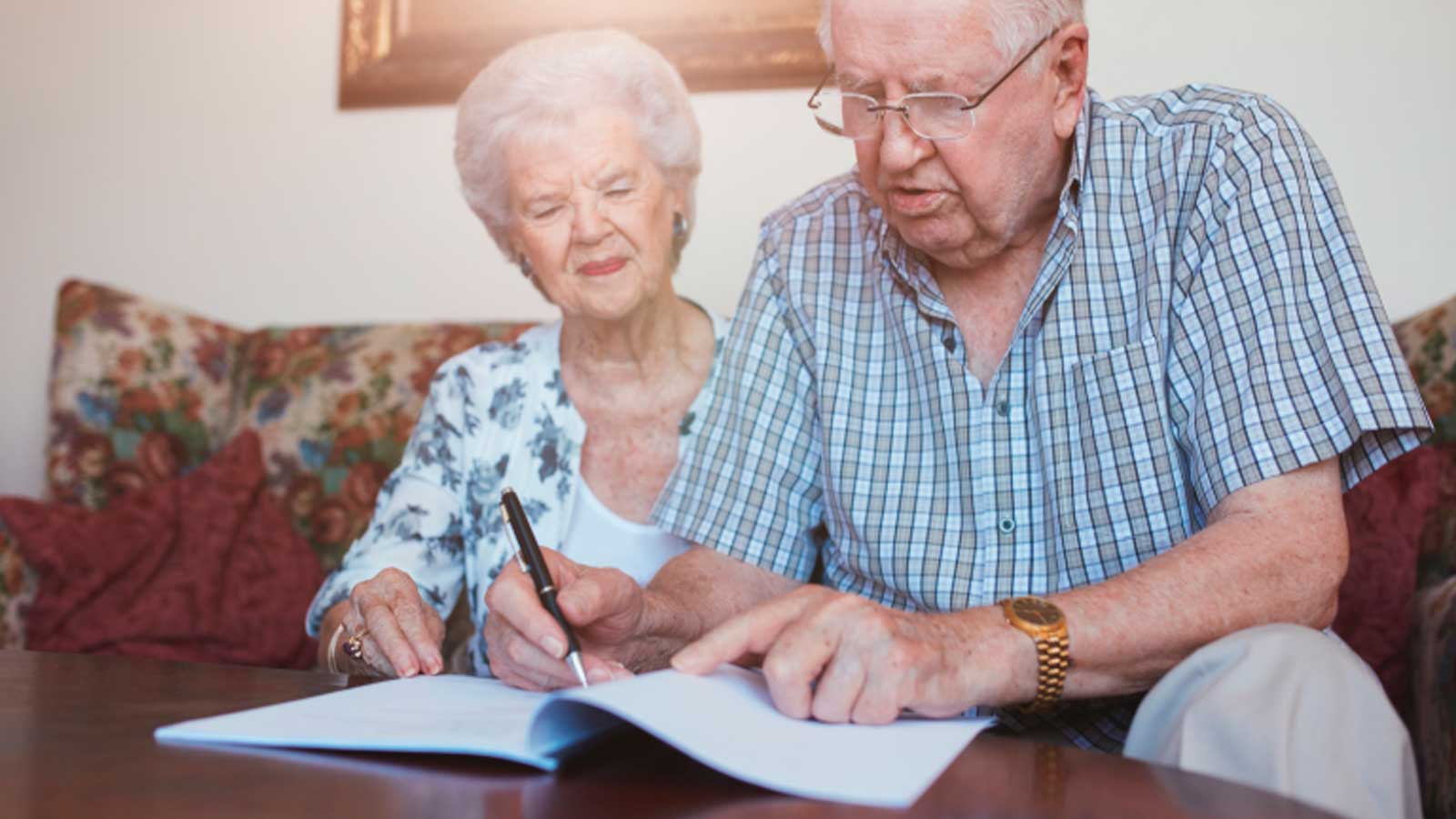 Dos personas mayores revisan y firman documentos en una mesa de su vivienda.