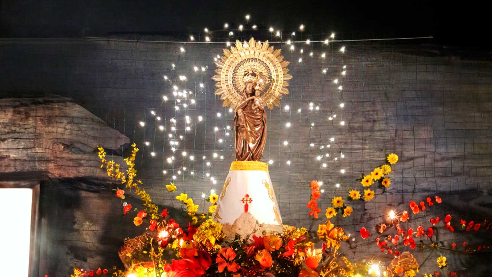 Imagen de la Virgen del Pilar iluminada y rodeada de flores durante una celebración religiosa.